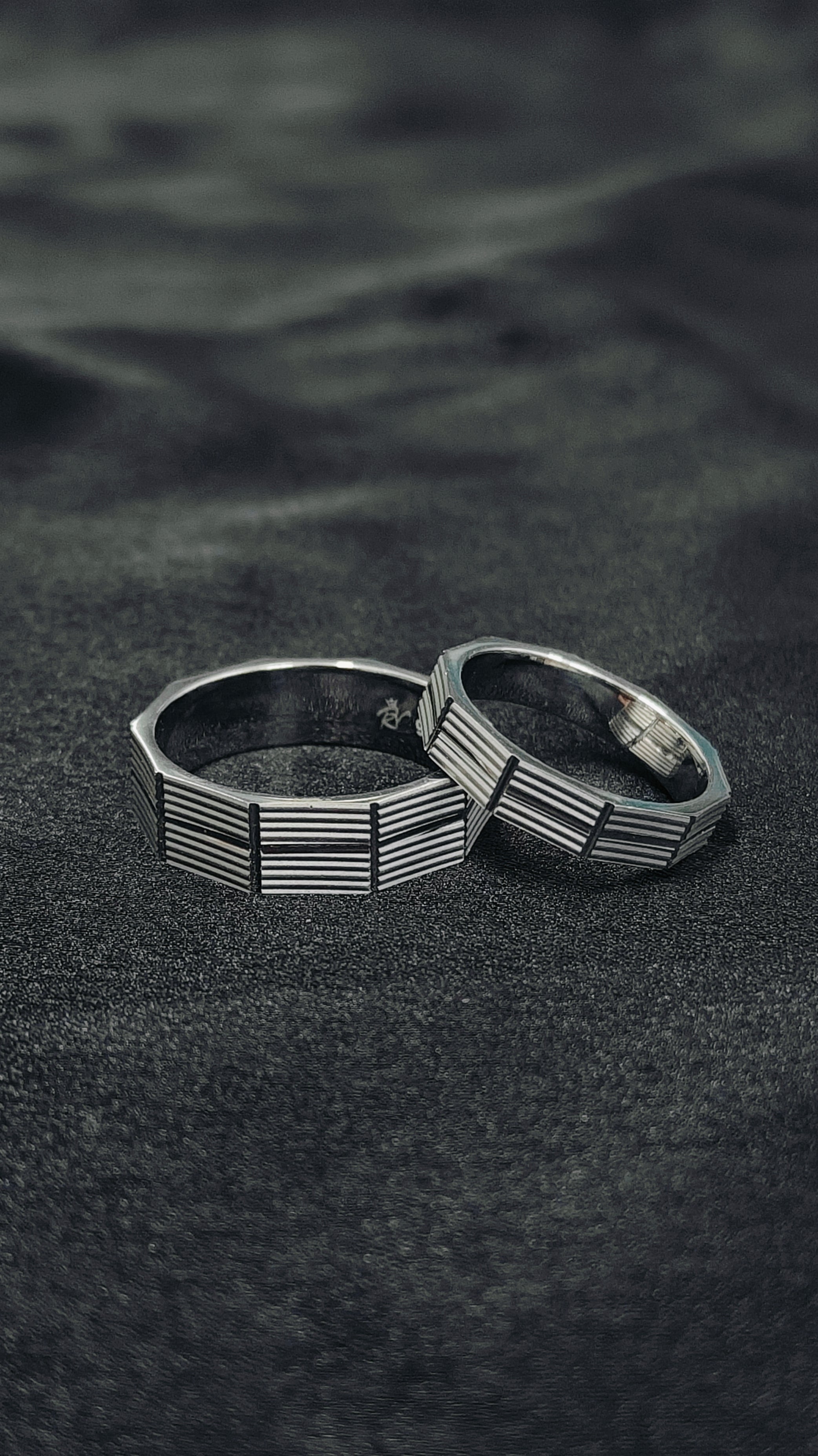 Two silver rings with geometric designs on a dark textured surface