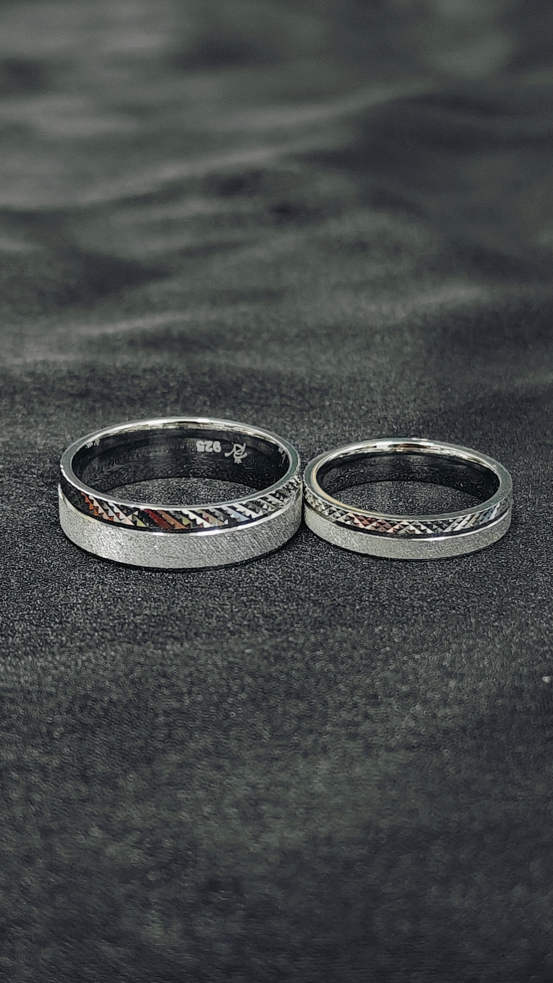 Two silver rings with intricate designs on a dark sandy surface.